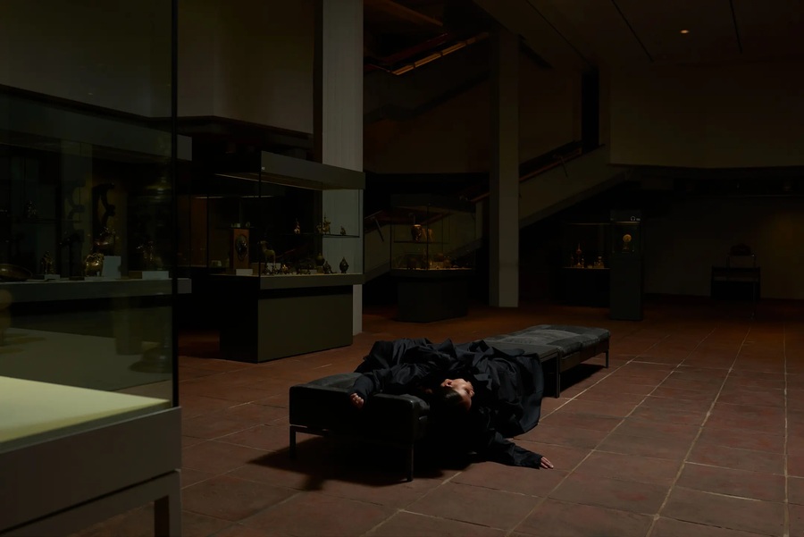 Figure draped in black fabric lying across benches inside the Kunstgewerbemuseum