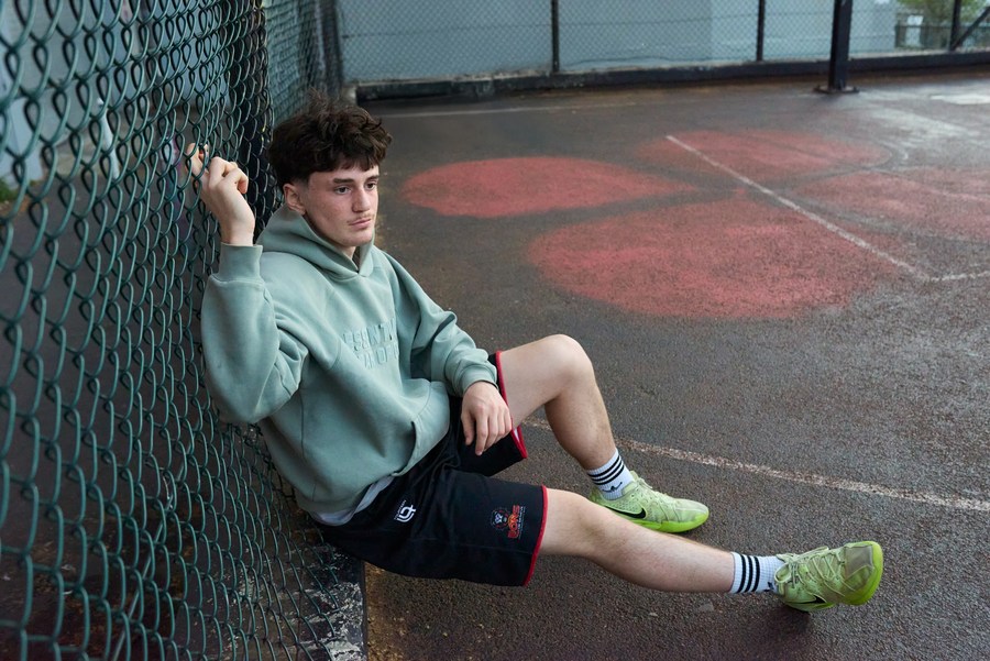 Seated figure in a pale green hoodie beside the court fence